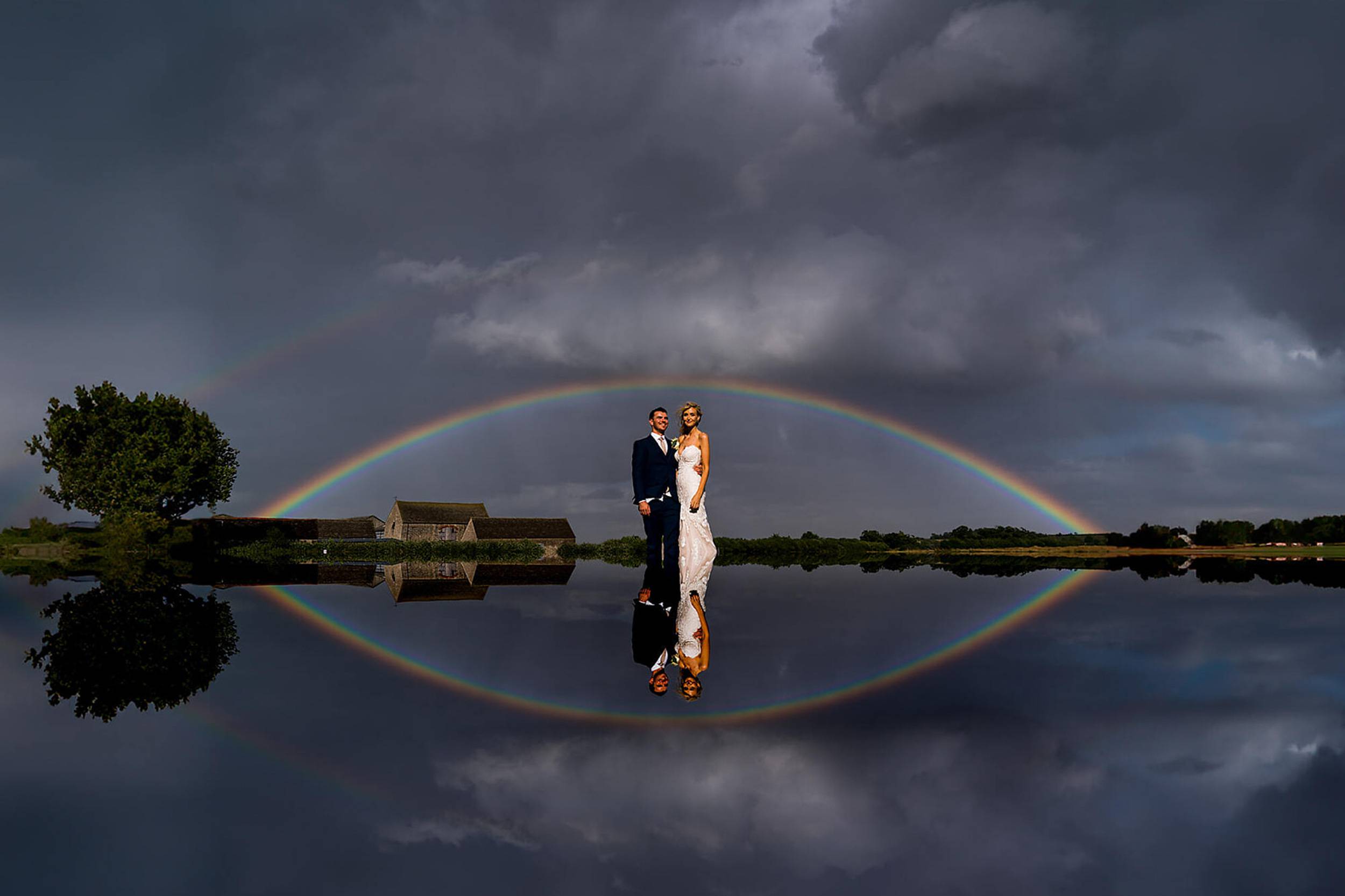 Bride and groom stand under a full rainbow and storm clouds, the whole scene mirrored in still water.
